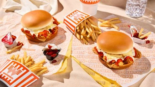 Two chicken sandwiches and french fries are displayed on branded wrapping paper, alongside condiments and a glass of water.