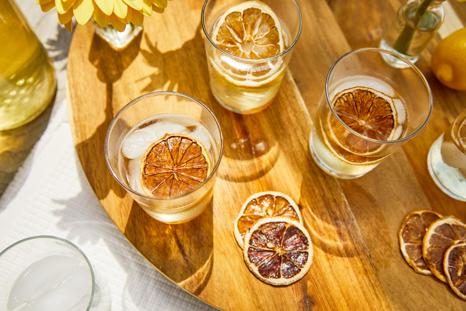 Overhead shot of three glasses of iced drinks with dehydrated lemon slices, arranged on wooden surface with flowers and soft lighting.