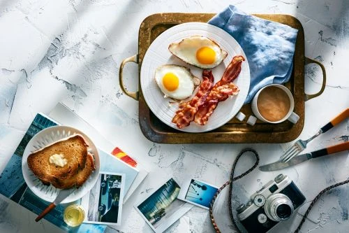 Overhead shot of breakfast featuring fried eggs, bacon, and toast on a tray, accompanied by coffee and a vintage camera for a nostalgic feel.