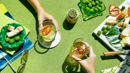 Overhead shot of two hands holding cocktails on a green tablecloth surrounded by plates of olives, bread, and cheese snacks.