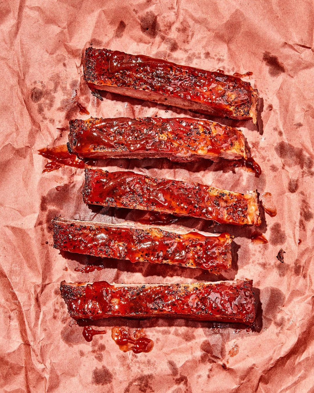 Overhead shot of five BBQ ribs with sauce, arranged on crumpled pink paper, highlighting texture and color for food photography.