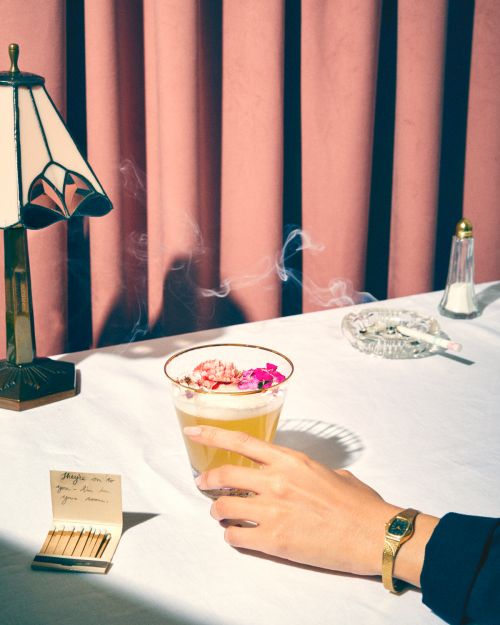 A hand holding a cocktail glass with flowers, with a lamp, salt shaker, cigarette, matches, and pink curtain in the background.