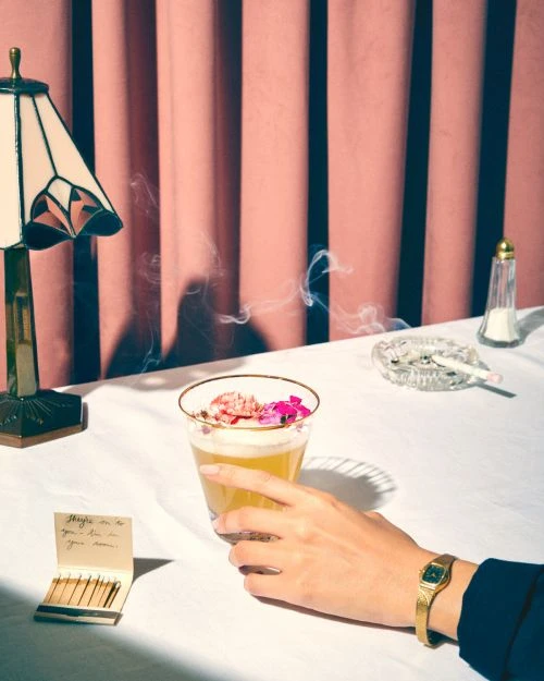 A hand holding a cocktail glass with flowers, with a lamp, salt shaker, cigarette, matches, and pink curtain in the background.