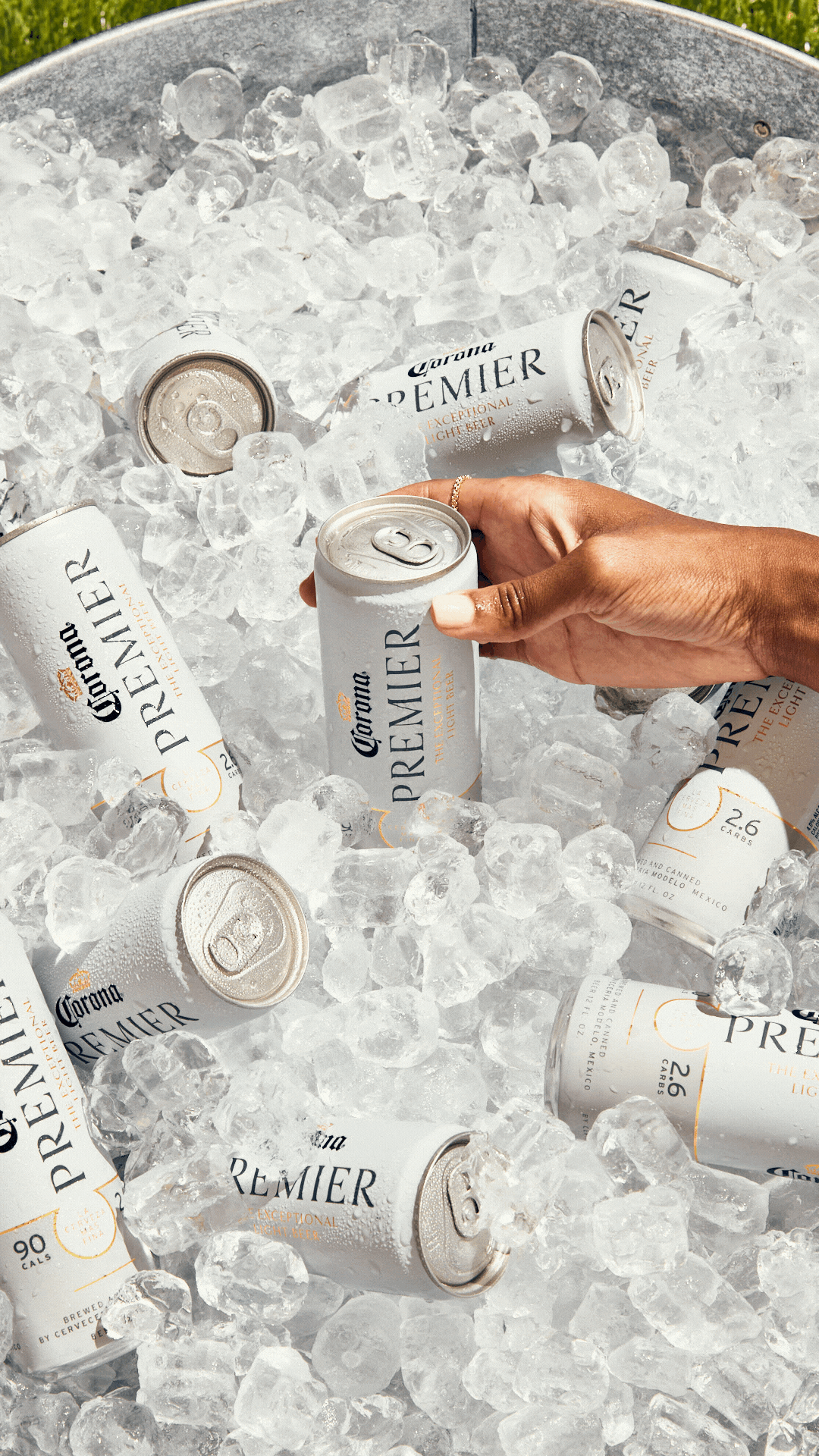 Several cans of Corona Premier beer are nestled in ice inside a metal bucket, with an arm reaching in.