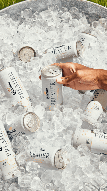 Several cans of Corona Premier beer are nestled in ice inside a metal bucket, with an arm reaching in.