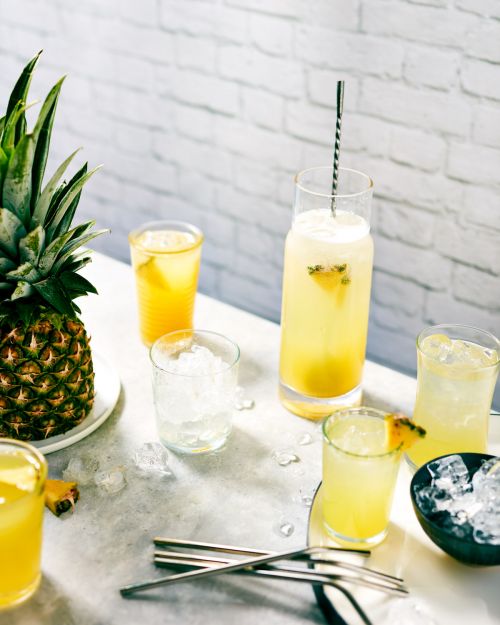 Pineapple cocktail assortment on a bright counter with ice, pineapple garnish, and a white brick background. Food and beverage photography.