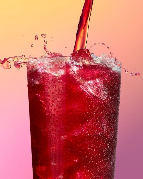 Close-up of a glass filled with a red beverage and ice, with more of the same drink being poured in, creating a splash against a pink background.