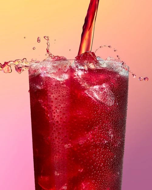 Close-up of a glass filled with a red beverage and ice, with more of the same drink being poured in, creating a splash against a pink background.