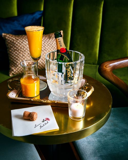 Still life of a mimosa drink, champagne bottle, and lit candle on a gold table with a velvet green background and white napkin.