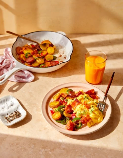 Breakfast plate features scrambled eggs, potatoes, bacon, jalapenos. Orange juice, skillet, salt are on a sunlit kitchen table.