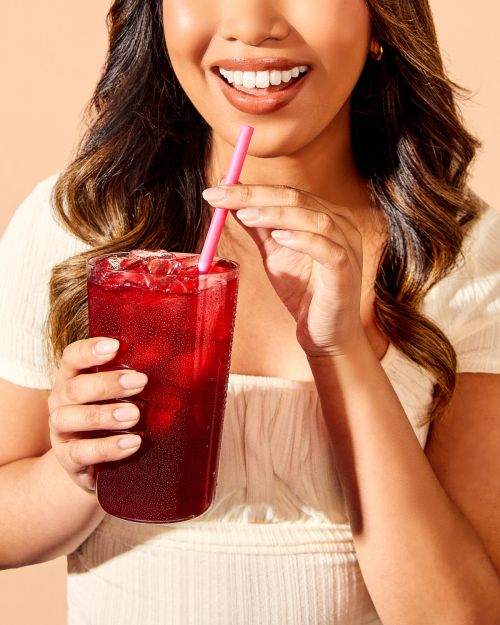 Woman smiling while holding a tall glass of red beverage with ice and a pink straw in an indoor environment, close-up shot.