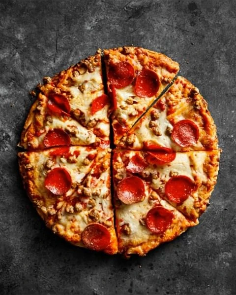 Overhead shot of a pepperoni and sausage pizza cut into slices on a dark textured surface. The pizza has a golden crust.