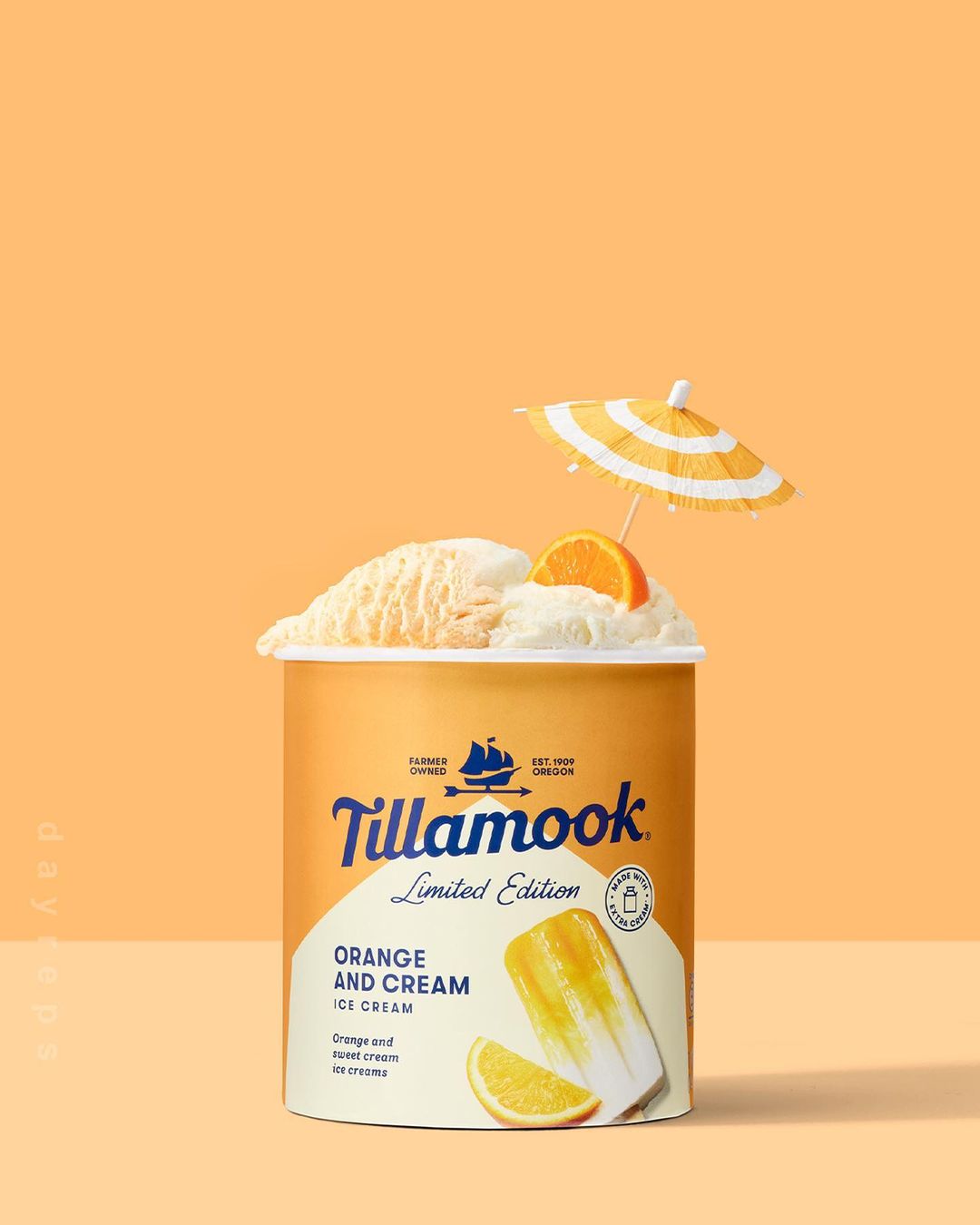 A container of Tillamook Orange and Cream ice cream sits against a soft orange backdrop, topped with ice cream, an orange slice, and a tiny umbrella.