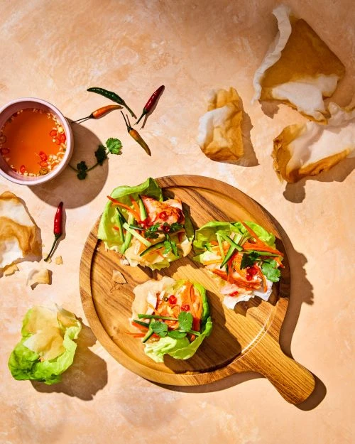 Overhead shot of three lettuce wraps with shrimp and vegetable filling, served on a round wooden board with dipping sauce.