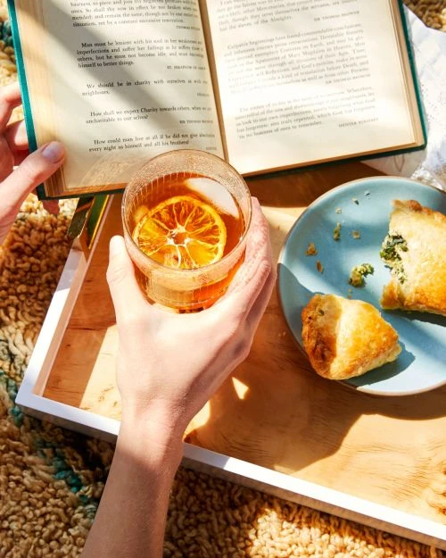Close-up: book and beverage on tray held by person. Drink has orange slice. Pastry on blue plate, patterned textile background.