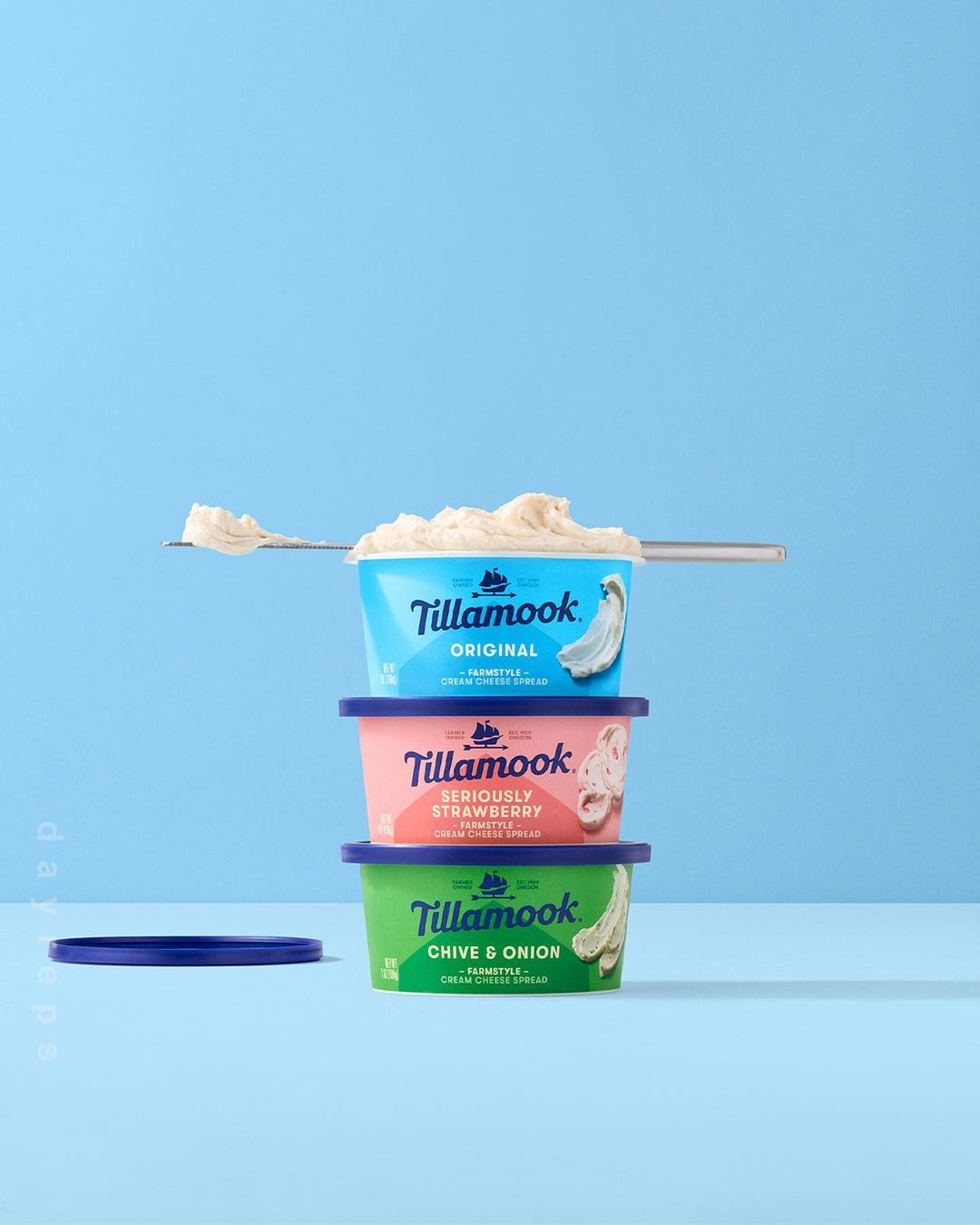 Stack of three colorful Tillamook cream cheese spread containers with spoon of cream cheese on light blue background in studio shot.