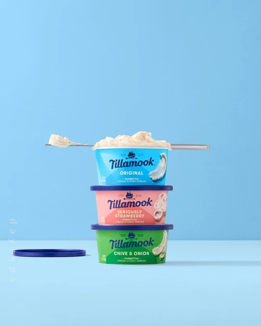 Stack of three colorful Tillamook cream cheese spread containers with spoon of cream cheese on light blue background in studio shot.