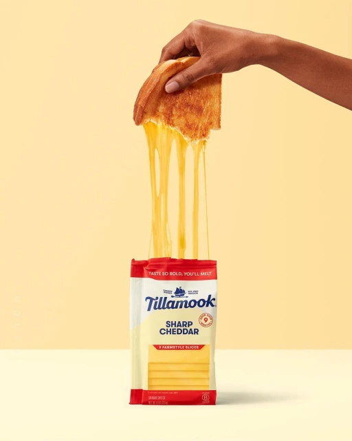 Hand holding grilled cheese sandwich with melting cheddar cheese, set against a yellow background with cheese slice packaging.