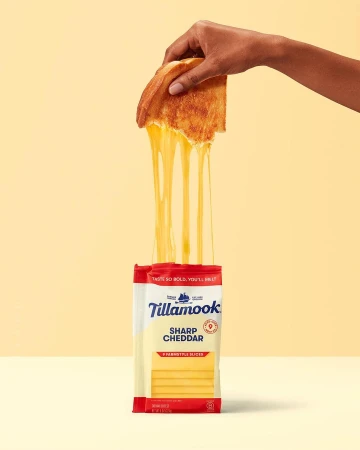 Hand holding grilled cheese sandwich with melting cheddar cheese, set against a yellow background with cheese slice packaging.