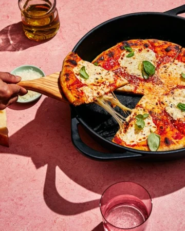 Overhead shot of a pizza being served from a skillet with a wooden spatula, adorned with basil leaves on a pink surface.
