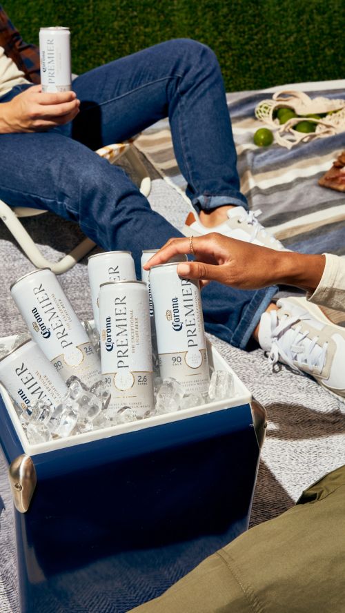 Outdoor picnic with Corona Premier cans in an ice cooler, with two people relaxing on a blanket in the background.
