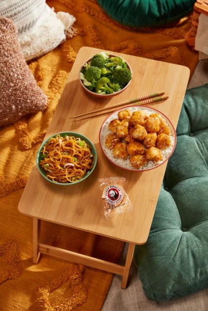 Overhead shot of a bamboo tray on cushions, holding Chinese food bowls with noodles, broccoli, chicken, rice, and chopsticks.