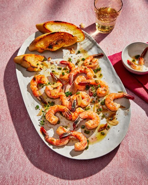 Overhead shot of grilled shrimp with herbs and garlic on a white platter, served with slices of toasted baguette and white wine.