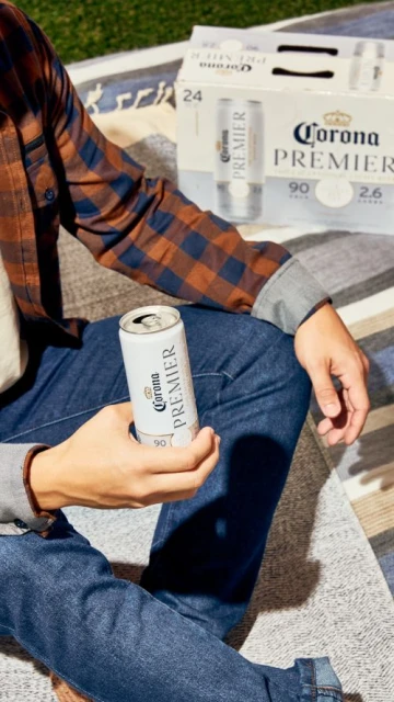 Adult male sitting with a can of Corona Premier beer in hand outdoors, with a carton of cans visible on a striped blanket.