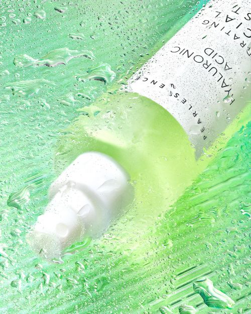 Close-up studio shot of a white bottle of hyaluronic acid serum product surrounded by water droplets against a green backdrop.