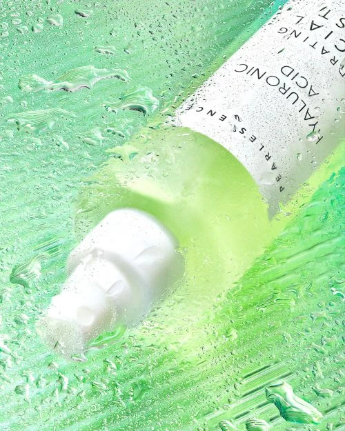 Close-up studio shot of a white bottle of hyaluronic acid serum product surrounded by water droplets against a green backdrop.