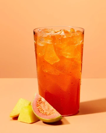 Refreshing guava juice in a clear glass with ice cubes, alongside slices of fresh guava fruit, set against a peach background.