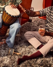 Two people sitting on a shaggy grey rug, one playing a djembe drum and another holding a glass of amber liquid