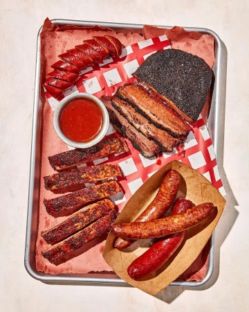 Overhead food photograph of Texas barbecue platter featuring brisket, ribs, sausage, and red sauce on butcher paper and metal tray