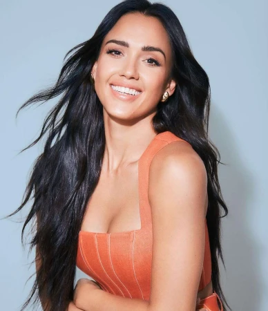 Close-up portrait of Jessica Alba with long dark hair, smiling and wearing an orange structured top against a light blue backdrop.