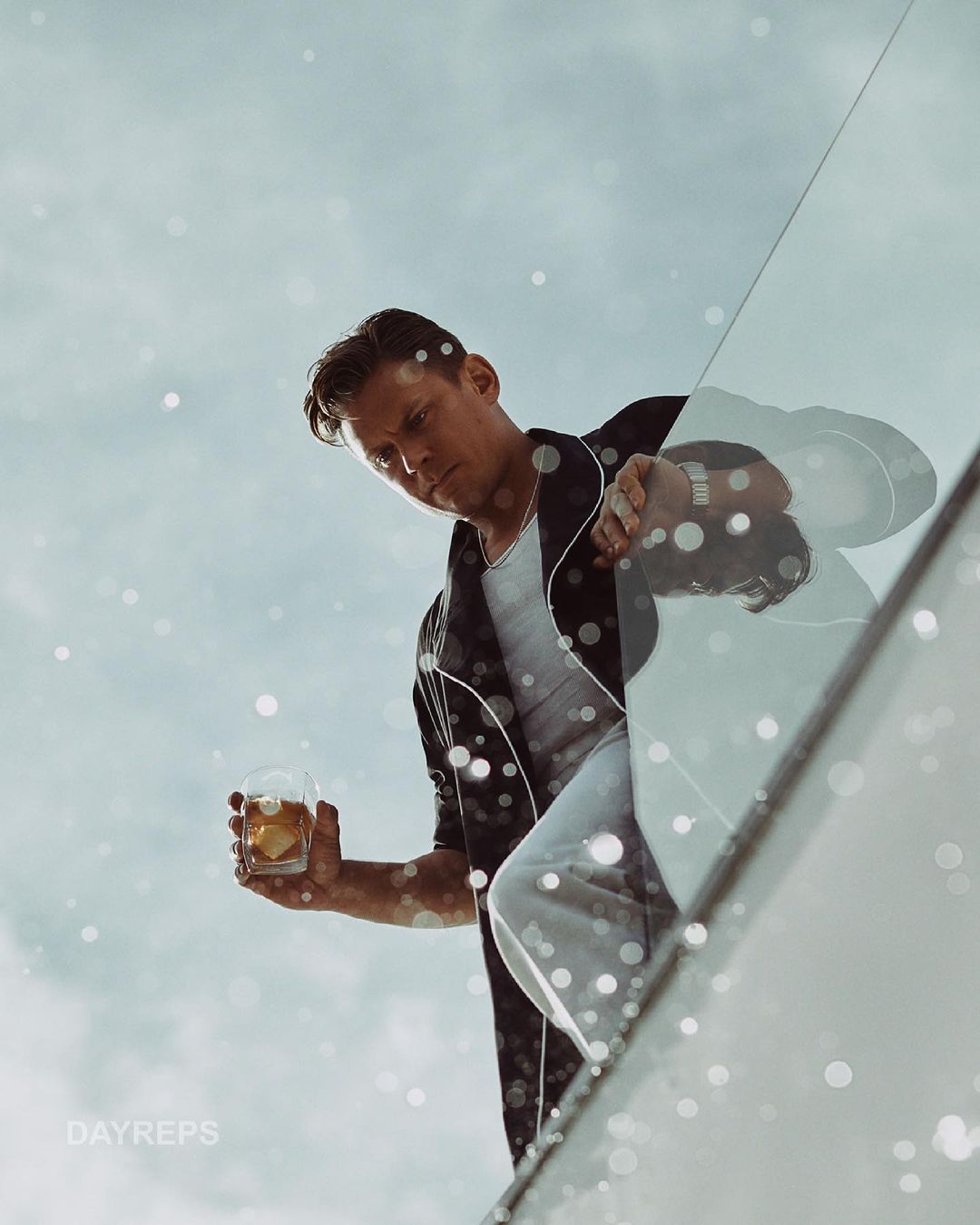Man in casual attire holding a cocktail glass, gazing downwards against a bright sky with bokeh effects and a modern architectural feature.