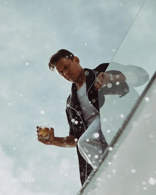 Man in casual attire holding a cocktail glass, gazing downwards against a bright sky with bokeh effects and a modern architectural feature.