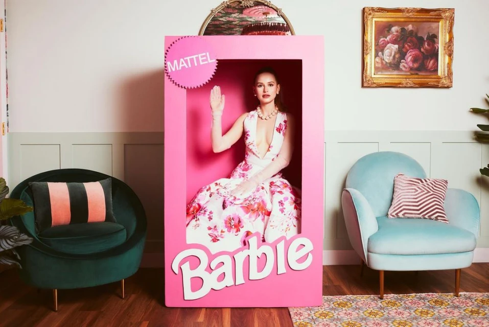 Woman in floral dress posing in a pink Barbie doll box in an indoor setting with vintage furniture and decor, creating a playful and nostalgic scene.