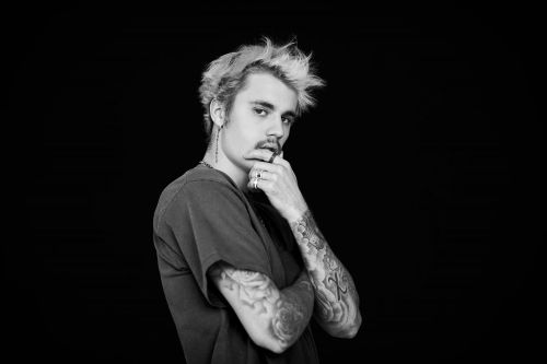 Monochrome portrait of Justin Bieber with blond spiky hair and arm tattoos posing in a dark t-shirt against a black backdrop.