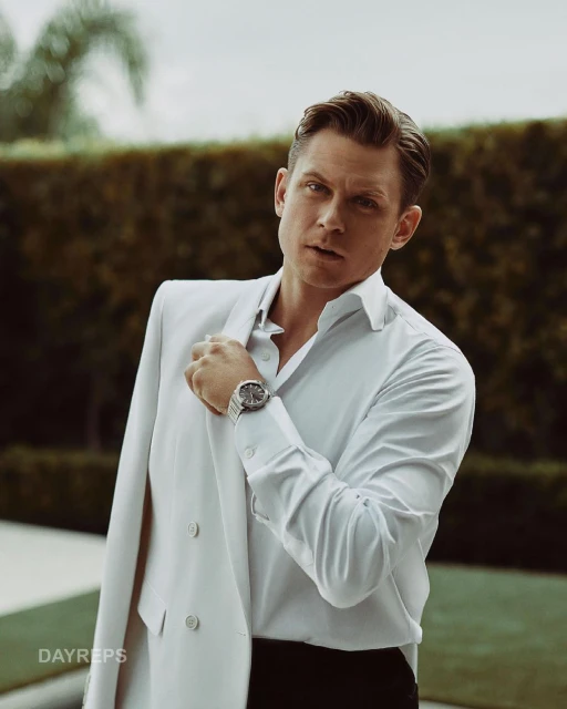 A man in a white suit and shirt poses outdoors with a wristwatch. The background features green foliage and soft, natural light.