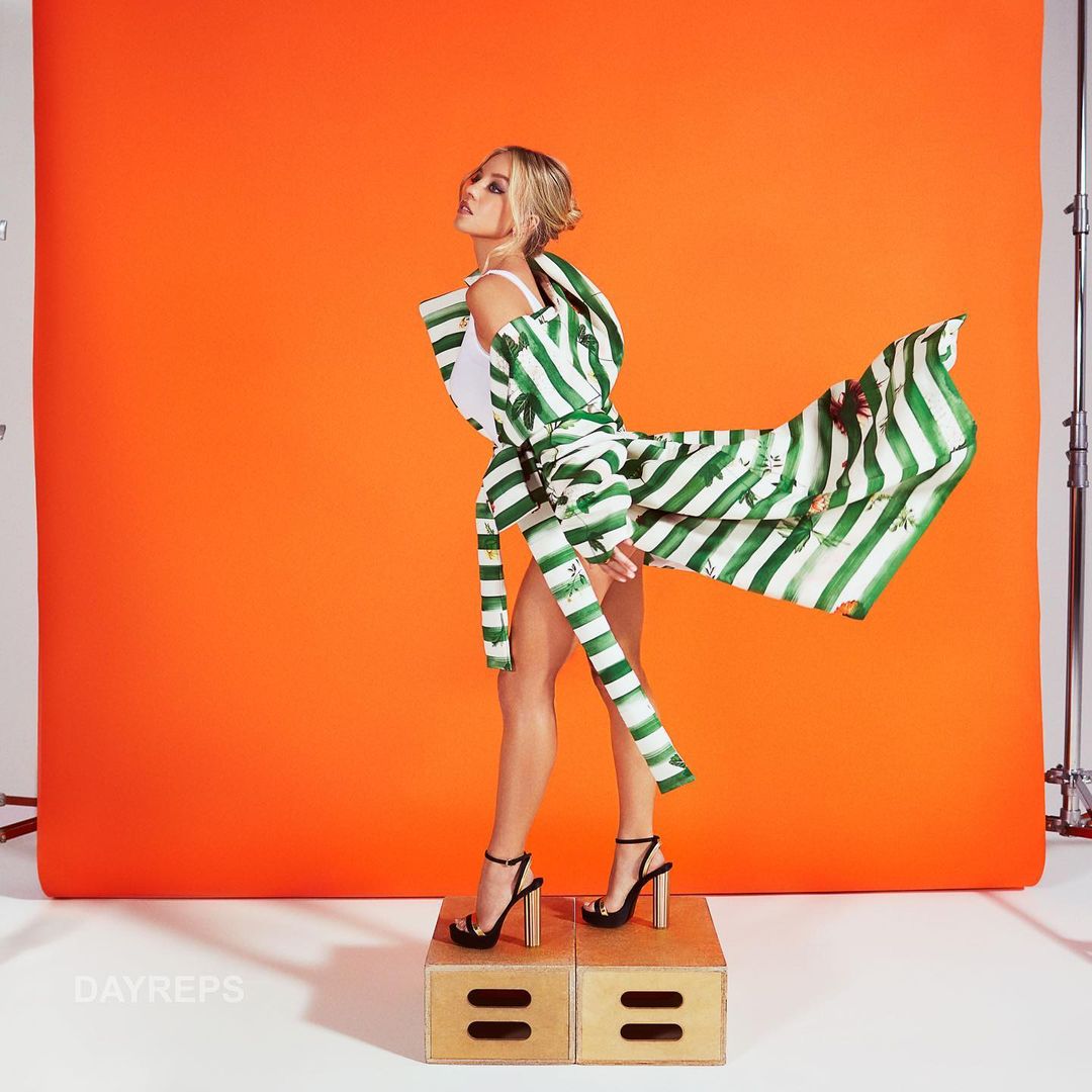 Fashion shot of a slender woman posing in a flowing green and white striped coat and black high heels against a bright orange background.