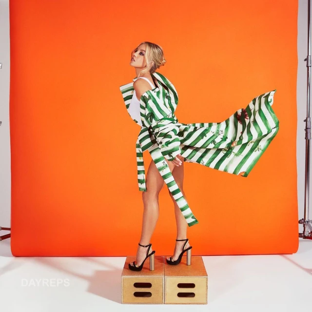 Fashion shot of a slender woman posing in a flowing green and white striped coat and black high heels against a bright orange background.