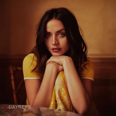 Ana de Armas leaning forward with her chin resting on her hands, wearing a yellow ribbed top against a warm interior background.