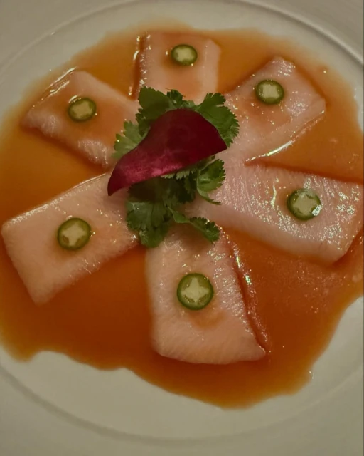 Overhead shot of hamachi crudo, featuring thin slices of fish topped with sliced jalapeños and cilantro in a light orange sauce.