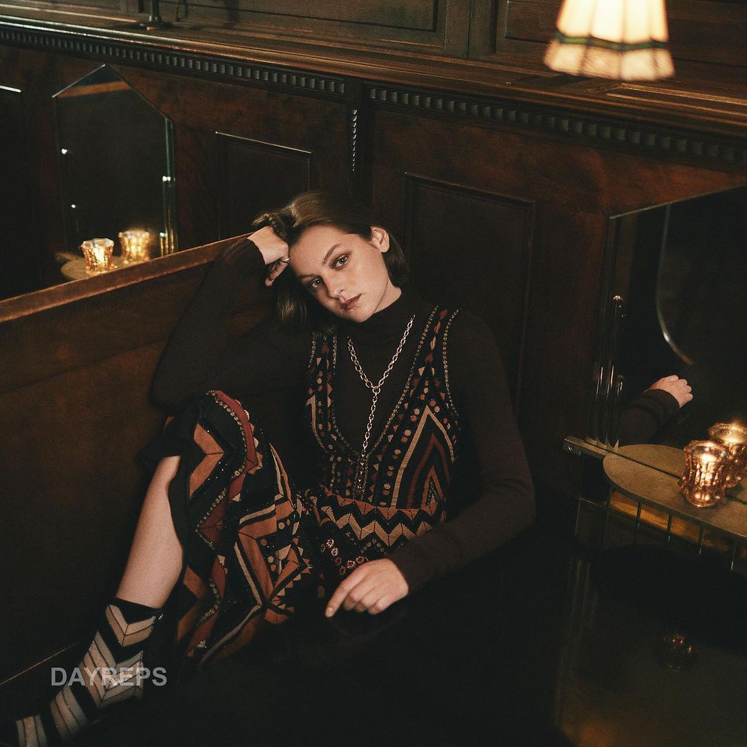 Stylish young woman with short hair poses indoors, wearing a printed vest over a turtleneck, accessorized with a silver chain necklace.