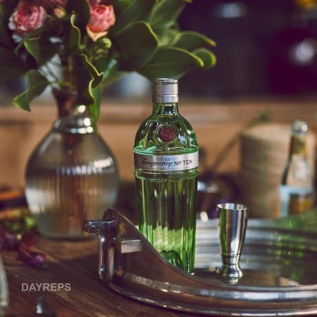 A green Tanqueray No. TEN gin bottle stands on a wooden table next to a silver jigger and floral arrangement. Selective focus shot.