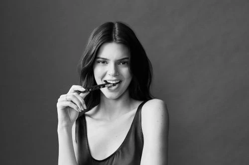 Black-and-white portrait of a smiling adult woman holding a black pen in her mouth, set against a plain gray studio backdrop.