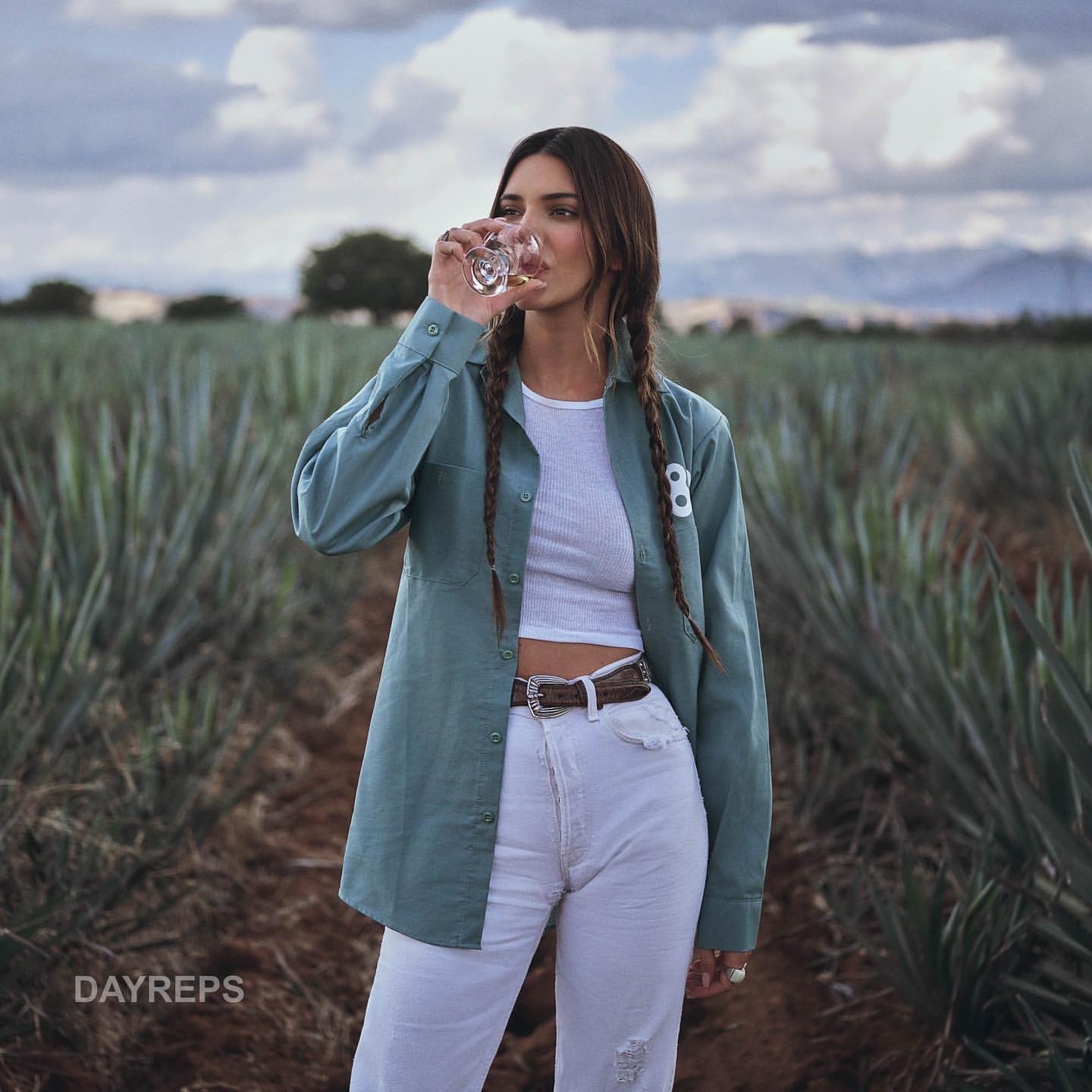 A woman is standing in a field of agave plants, wearing a teal shirt, white top and pants, holding a glass of clear liquid.