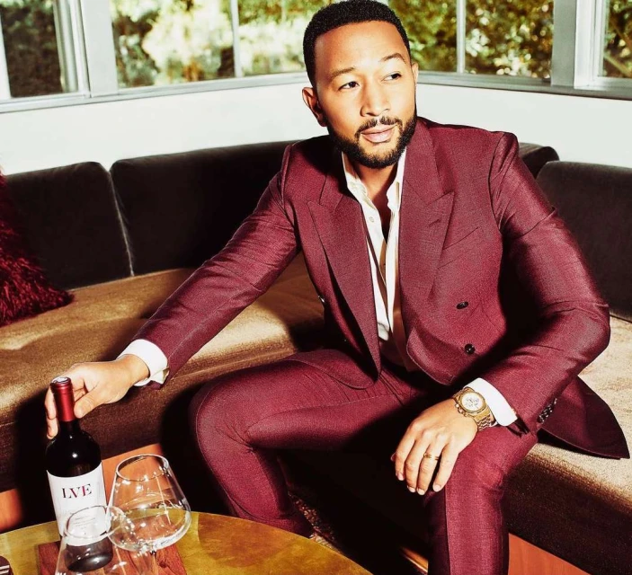 John Legend poses indoors with a wine bottle and glasses, wearing a suit. He sits on a velvet couch with a coffee table.