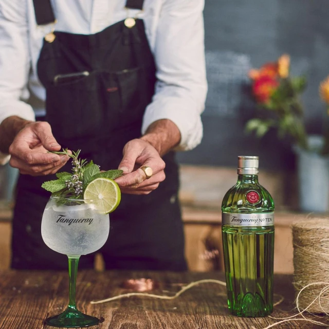Close-up shot of an adult preparing a Tanqueray gin cocktail with lime, mint, and herbs on a wooden table with a bottle.