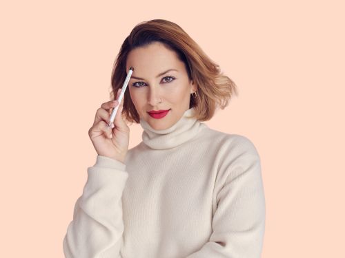 Woman with short brown hair and red lipstick holds a white pencil near her forehead, posing in front of a peach background.